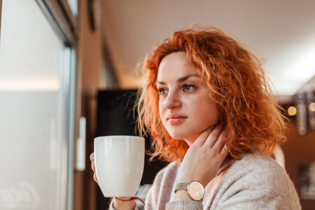 Red-haired curly young bright woman with green eyes drinks hot coffee in cozy cafe. Beautiful model in stylish clothes. Europe.の写真素材