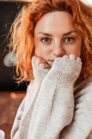 Red-haired curly young bright woman with green eyes drinks hot coffee in cozy cafe. Beautiful model in stylish clothes.の写真素材