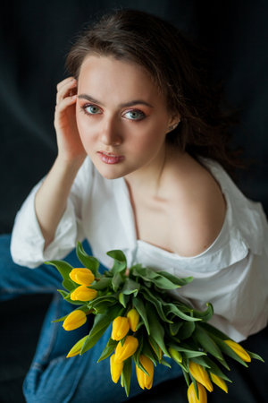 Spring portrait of young beautiful woman with blue eyes and curly hair with bouquet of yellow tulips on black background. Happy model in jeans and white shirt.の写真素材