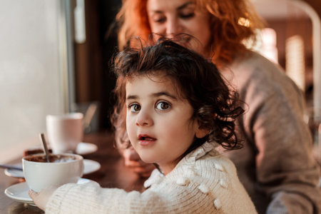 Beautiful young red-haired mother with cute curly-haired daughter are sitting in cozy cafe and drinking hot school. mother's day.の写真素材