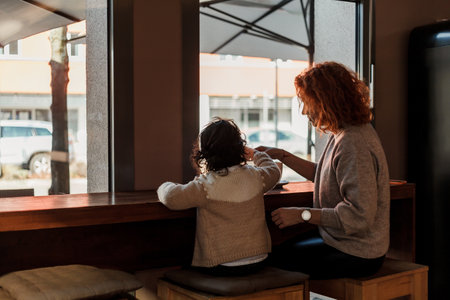 Beautiful young red-haired mother with cute curly-haired daughter are sitting in cozy cafe and drinking hot school. mother's day.の写真素材