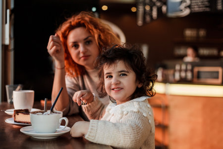 Beautiful young red-haired mother with cute curly-haired daughter are sitting in cozy cafe and drinking hot school. mother's day.の写真素材