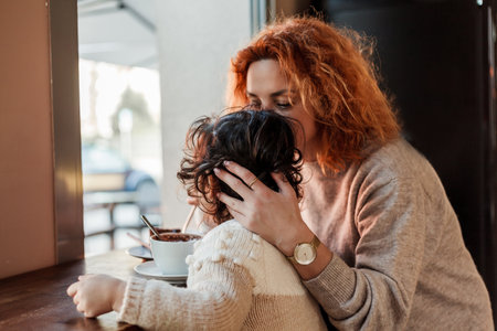Beautiful young red-haired mother with cute curly-haired daughter are sitting in cozy cafe and drinking hot school. mother's day.の写真素材