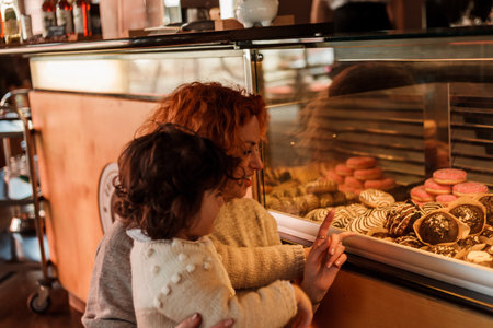 Cute little curly girl 3 years old with a young red-haired mother in the bakery at the counter chooses pastries. cafe. Cozy.の写真素材