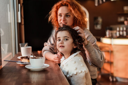 Beautiful young red-haired mother with cute curly-haired daughter are sitting in cozy cafe and drinking hot school. mother's day.の写真素材