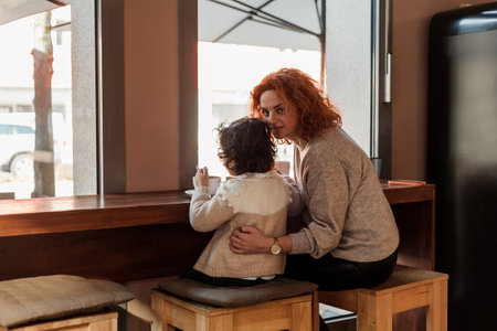 Beautiful young red-haired mother with cute curly-haired daughter are sitting in cozy cafe and drinking hot school. mother's day.の写真素材