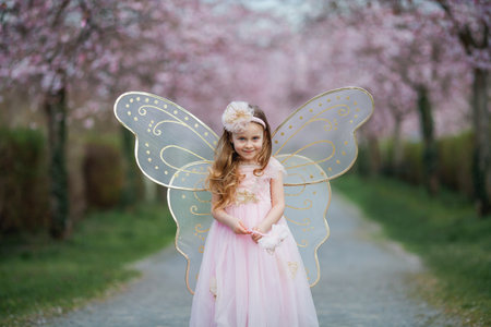 Cute little long-haired curly-haired girl in a beautiful pink fairy costume with big wings in a blooming sakura garden. Spring.の写真素材