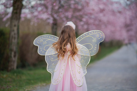 Cute little long-haired curly-haired girl in a beautiful pink fairy costume with big wings in a blooming sakura garden. Spring.の写真素材