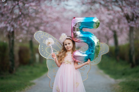 Cute little long-haired curly-haired girl in a beautiful pink fairy costume with big wings in a blooming sakura garden. Spring.の写真素材