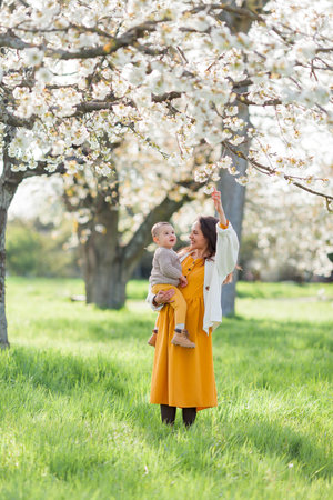 Happy young mother with little son walks in blooming garden. Mom with 1 year old baby.の写真素材