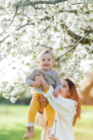 Happy young mother with little son walks in blooming garden. Mom with 1 year old baby.の写真素材