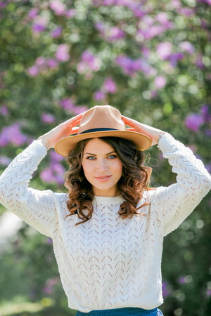 Portrait of beautiful young dark-haired woman with blue eyes in hat near the blooming. Happy model with curly hair.の写真素材