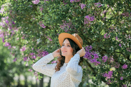 Portrait of beautiful young dark-haired woman with blue eyes in hat near the blooming. Happy model with curly hair.の写真素材
