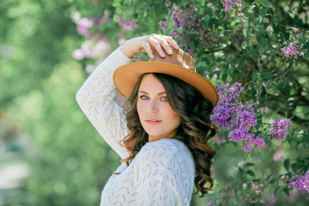 Portrait of beautiful young dark-haired woman with blue eyes in hat near the blooming. Happy model with curly hair.の写真素材