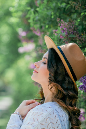 Portrait of beautiful young dark-haired woman with blue eyes in hat near the blooming. Happy model with curly hair.の写真素材