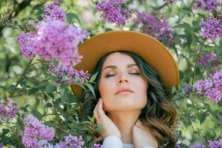 Portrait of beautiful young dark-haired woman with blue eyes in hat near the blooming. Happy model with curly hair.の写真素材