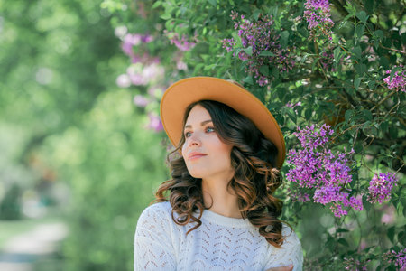 Portrait of beautiful young dark-haired woman with blue eyes in hat near the blooming. Happy model with curly hair.の写真素材
