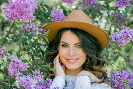 Portrait of beautiful young dark-haired woman with blue eyes in hat near the blooming. Happy model with curly hair.の写真素材