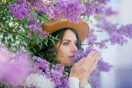 Portrait of beautiful young dark-haired woman with blue eyes in hat near the blooming. Happy model with curly hair.の写真素材