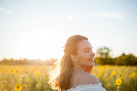 Young beautiful blonde woman in white dress in the field of sunflowers at sunset. Evening sun rays.