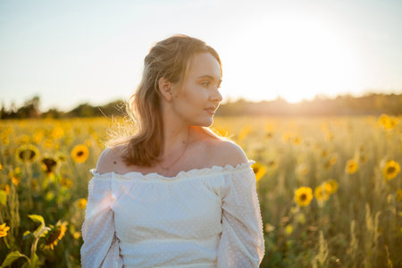 Young beautiful blonde woman in white dress in the field of sunflowers at sunset. Evening sun rays.の写真素材