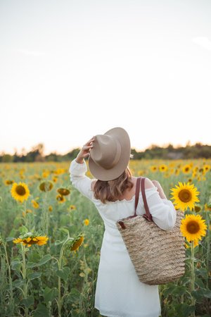 Portrait of beautiful young woman 33 years old in hat in sunflower field at sunset. Happy model in white dress on summer evening in nature.の写真素材