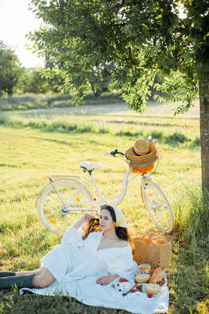 Beautiful young dark-haired woman on picnic in the field. Model in stylish clothes with bicycle.の写真素材