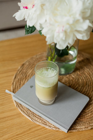 Home cozy interior. A bouquet of peonies in a glass vase, a glass of matchalate on a wooden coffee table.の写真素材