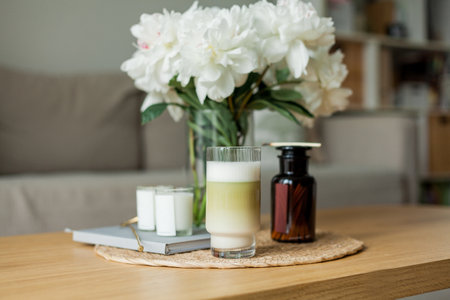 Home cozy interior. Candles, a bouquet of peonies in a glass vase, a glass of matchalate on a wooden coffee table.の写真素材