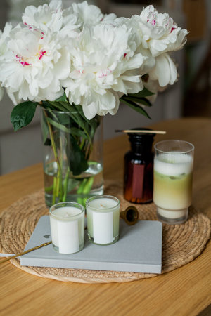 Home cozy interior. Candles, a bouquet of peonies in a glass vase, a glass of matchalate on a wooden coffee table.の写真素材