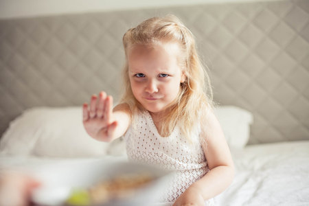Little cute white-haired girl eats granola on the bed in her bright room. Health food.の写真素材