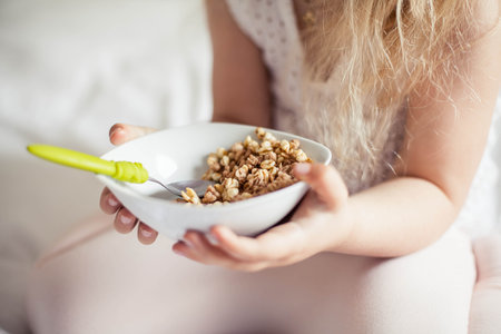 Little cute white-haired girl eats granola on the bed in her bright room. Health food.の写真素材