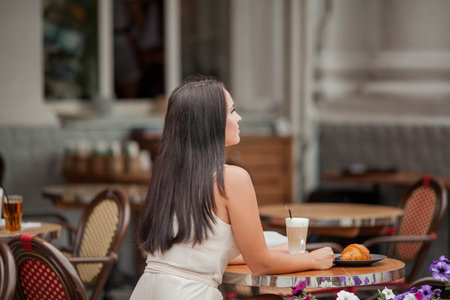 Young beautiful woman is sitting in a french cafe and drinking cappuccino, eating a croissant. bakery products. Summer.の写真素材
