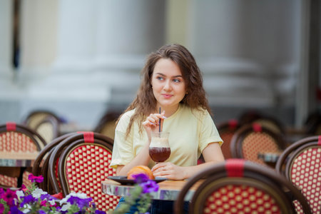 Beautiful dark-haired young curly woman sits in summer cafe and drinks coffee cocktail. warm.の写真素材