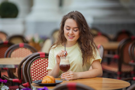 Beautiful dark-haired young curly woman sits in summer cafe and drinks coffee cocktail. warm.の写真素材