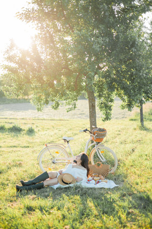 Beautiful young dark-haired woman on picnic in the field. Model in stylish clothes with bicycle.の写真素材