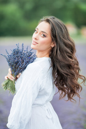 Lavender field. Beautiful dark-haired curly woman in white simple dress in a field of purple flowers.の写真素材