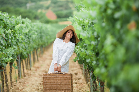 Beautiful young dark-haired girl in the vineyards. Stylish model with wicker hat and suitcase. Travel. Summer holidays. Trip.の写真素材
