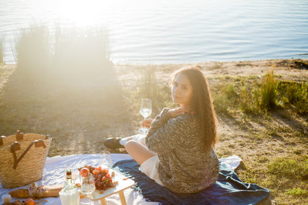 Beautiful young curly woman in warm cardigan on cozy autumn picnic by the lake with fruits, pastries and glass of white wine. Sun rays.の写真素材