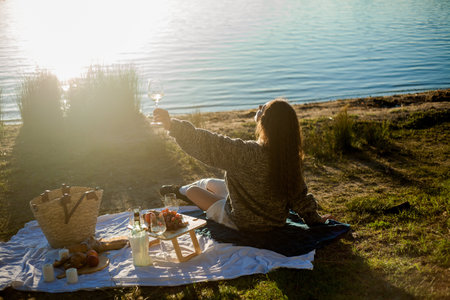 Beautiful young curly woman in warm cardigan on cozy autumn picnic by the lake with fruits, pastries and glass of white wine. Sun rays.の写真素材