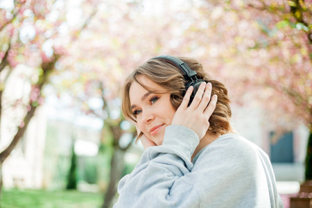 Beautiful young woman in comfortable clothes listens to music in headphones in blooming park. Sakura Pink flowers.の写真素材