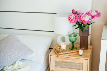 Bouquet of peonies in glass pink vase, lamp, candles on wooden stand. Bedroom.