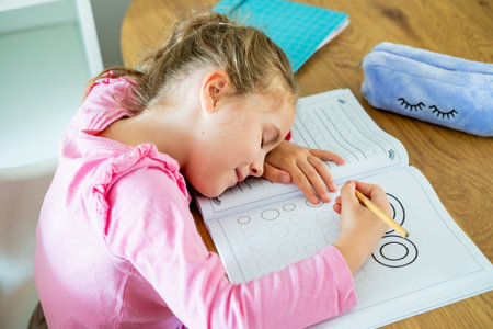 Cute little girl of 6 years old is doing homework at home in the kitchen at the table. Back to...