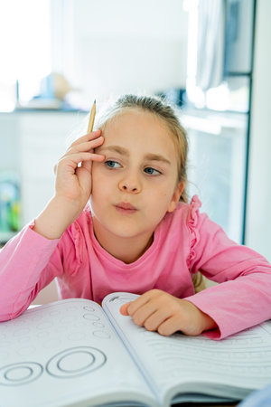 Cute little girl of 6 years old is doing homework at home in the kitchen at the table. Back to...