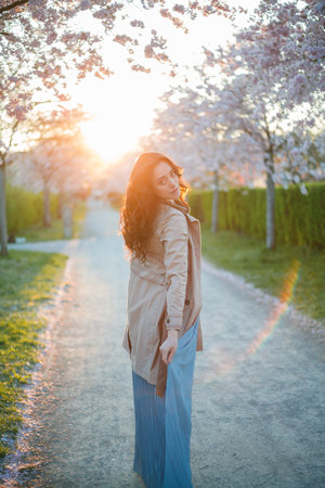 Young beautiful woman 36 years old with curly long dark hair near blooming sakura at sunset. Model in blue dress in spring in blooming park.の写真素材