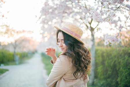 Young beautiful woman 36 years old with curly long dark hair near blooming sakura at sunset. Model in blue dress in spring in blooming park.の写真素材