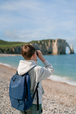Cute boy with blond hair, 10 years old, with sports backpack on the seashore in France, looks through binoculars at the horizon. Cliffs. Etretat.の写真素材