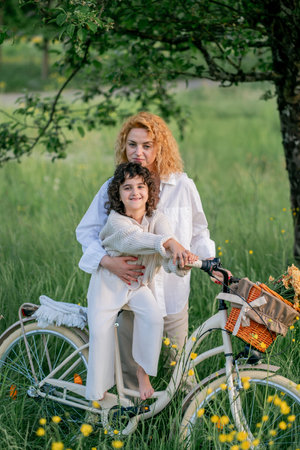 Mother and daughter enjoying a sunny day with a bicycle in a field of flowers. Red-haired young woman with a 5-year-old girl resting in nature.の写真素材