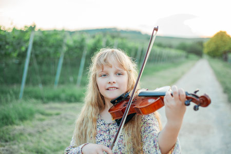 Closeup of a little girl with long blond hair playing the violin outdoor. Child holding a musical instrument in her hands. Concert in nature. Development.の写真素材