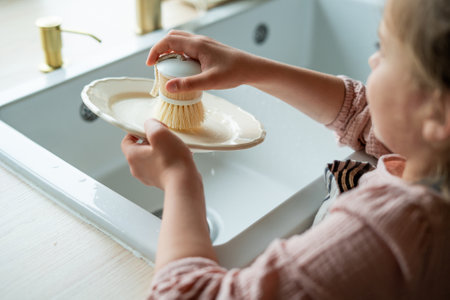 Closeup of children's hands washing a plate near the sink in the kitchen. Little girl holding an ecofriendly dishwashing brush.の写真素材